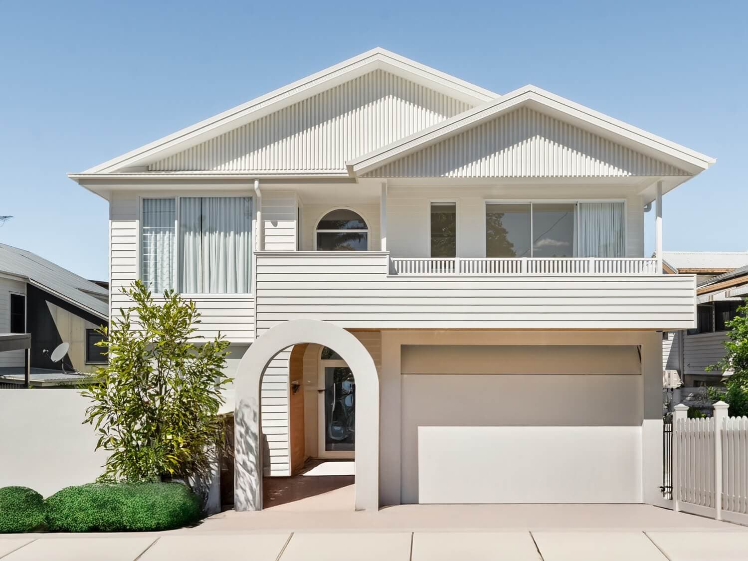 Exterior facade of a modern two-storey custom home featuring a striking arched entryway, white vertical cladding, and a classic white picket fence.