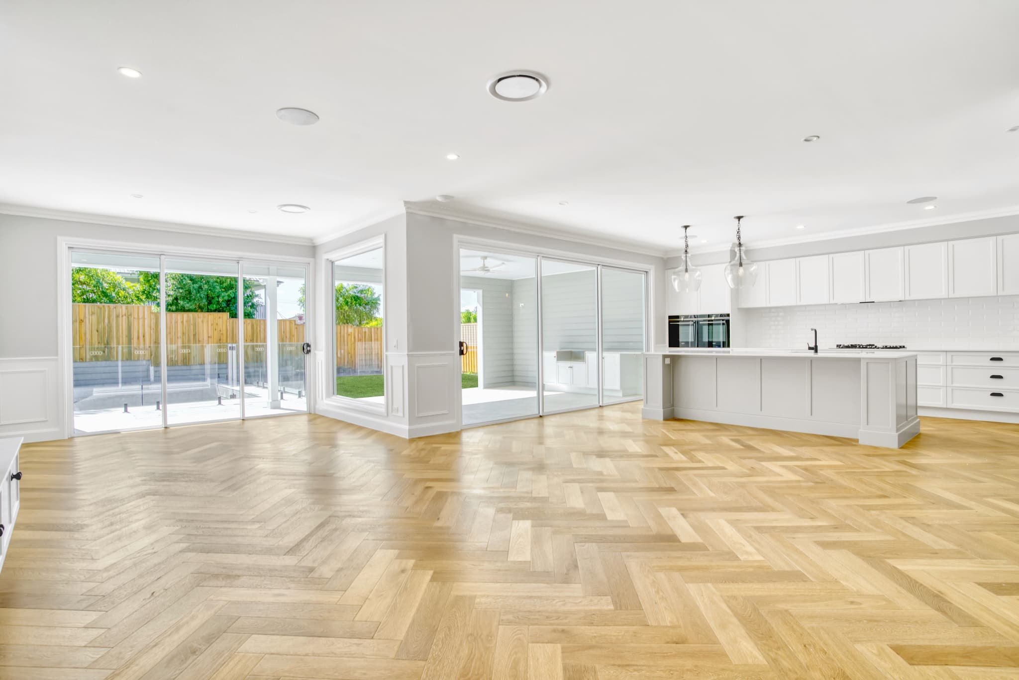 A luxury custom open plan home design in Brisbane featuring herringbone timber floors, a white shaker kitchen, and flush-threshold sliding doors opening to an alfresco pool area.