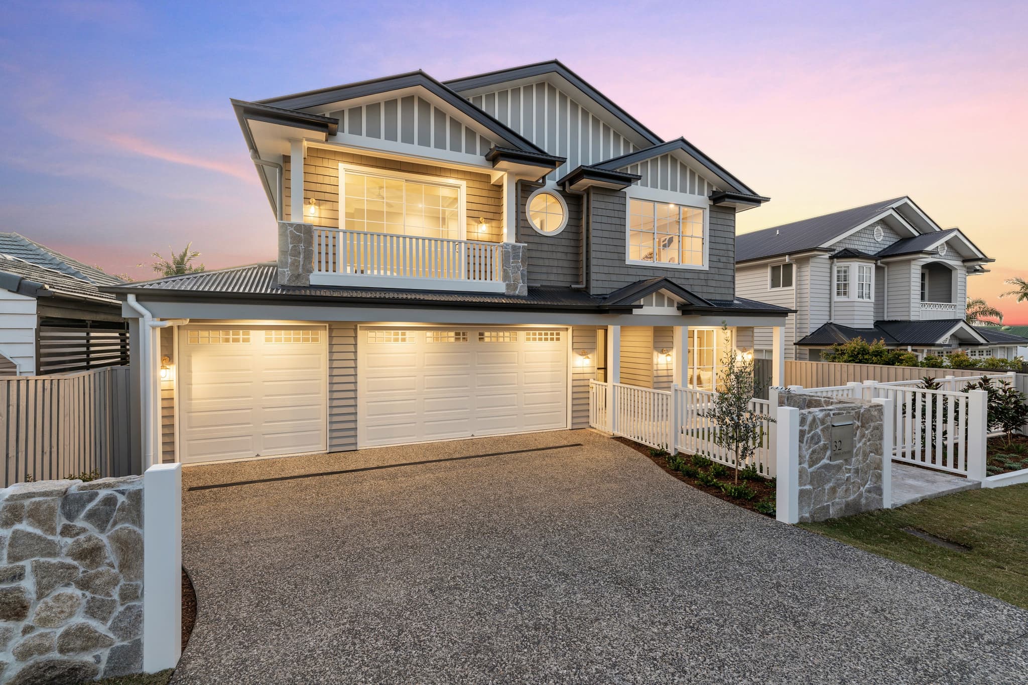 A custom home by premium Hampton style builders in Brisbane, featuring a classic gabled roof, weatherboards, and a stone fence at dusk.