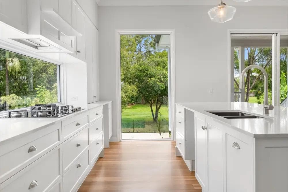 Kitchen with white shaker cabinetry opening to a leafy sloping backyard through full-height glazing.