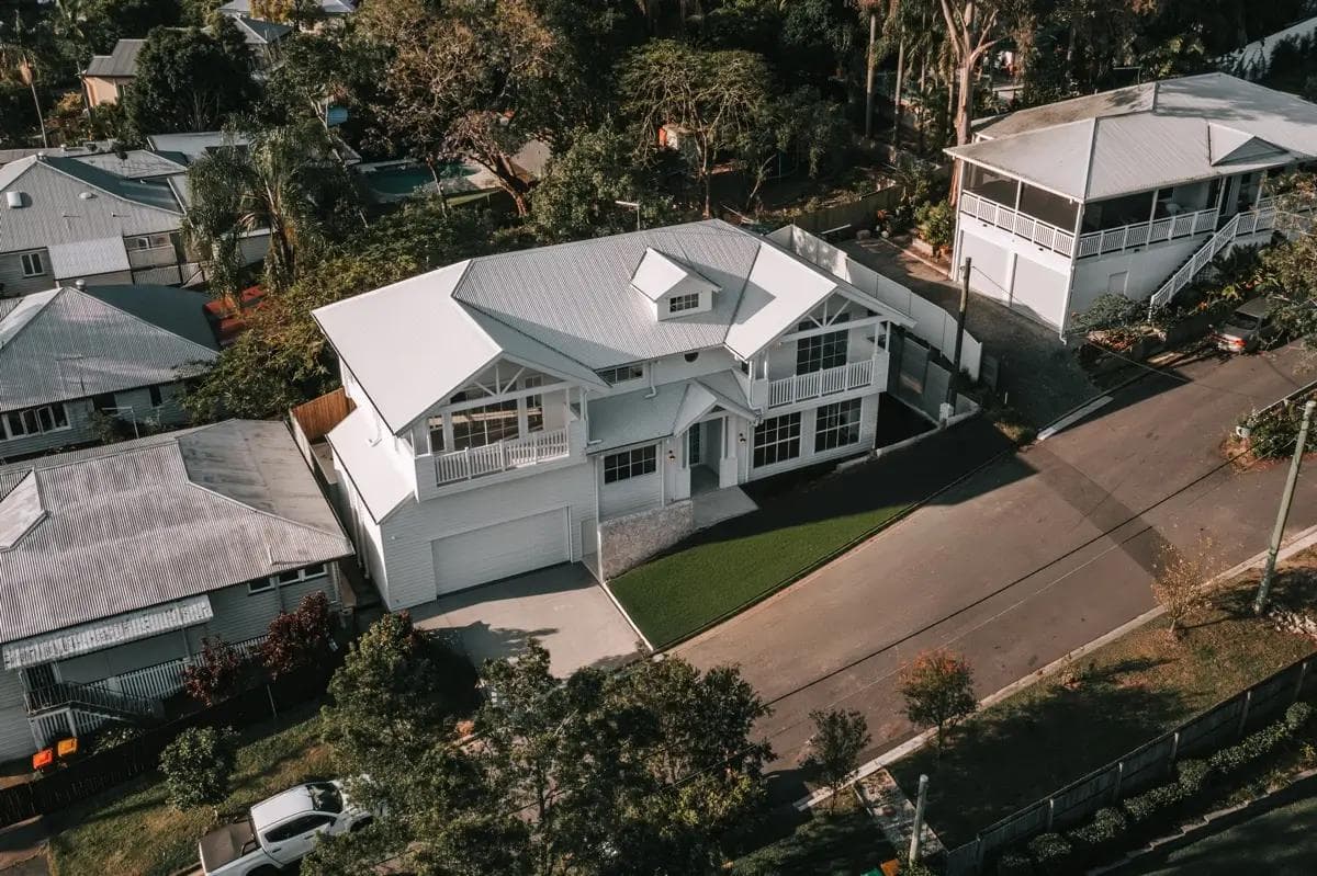 Aerial drone view of a large, custom-built two-storey white Hamptons-style home featuring gabled rooflines, front balconies, and a light metal roof, situated on a leafy suburban street.