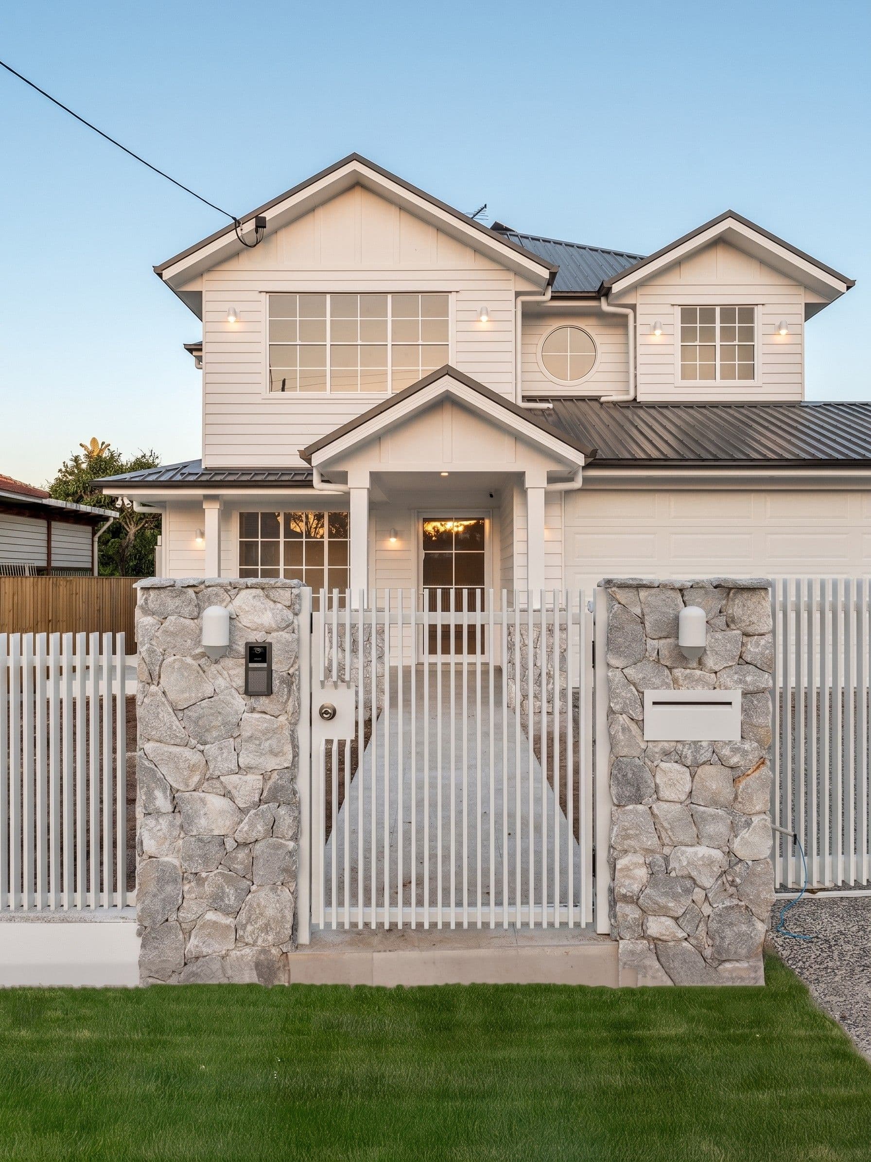 White Hamptons-style home in Mount Gravatt East with stone entry piers and picket fencing at dusk.