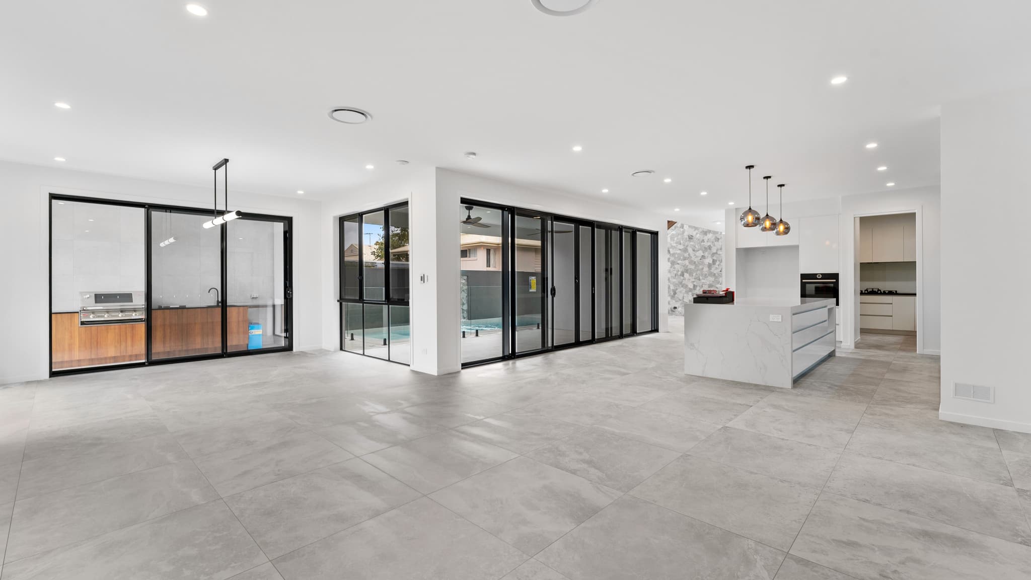 An expansive, open-plan living area and kitchen in a custom Brisbane home design, showcasing massive glass doors opening seamlessly to a swimming pool and outdoor entertaining area