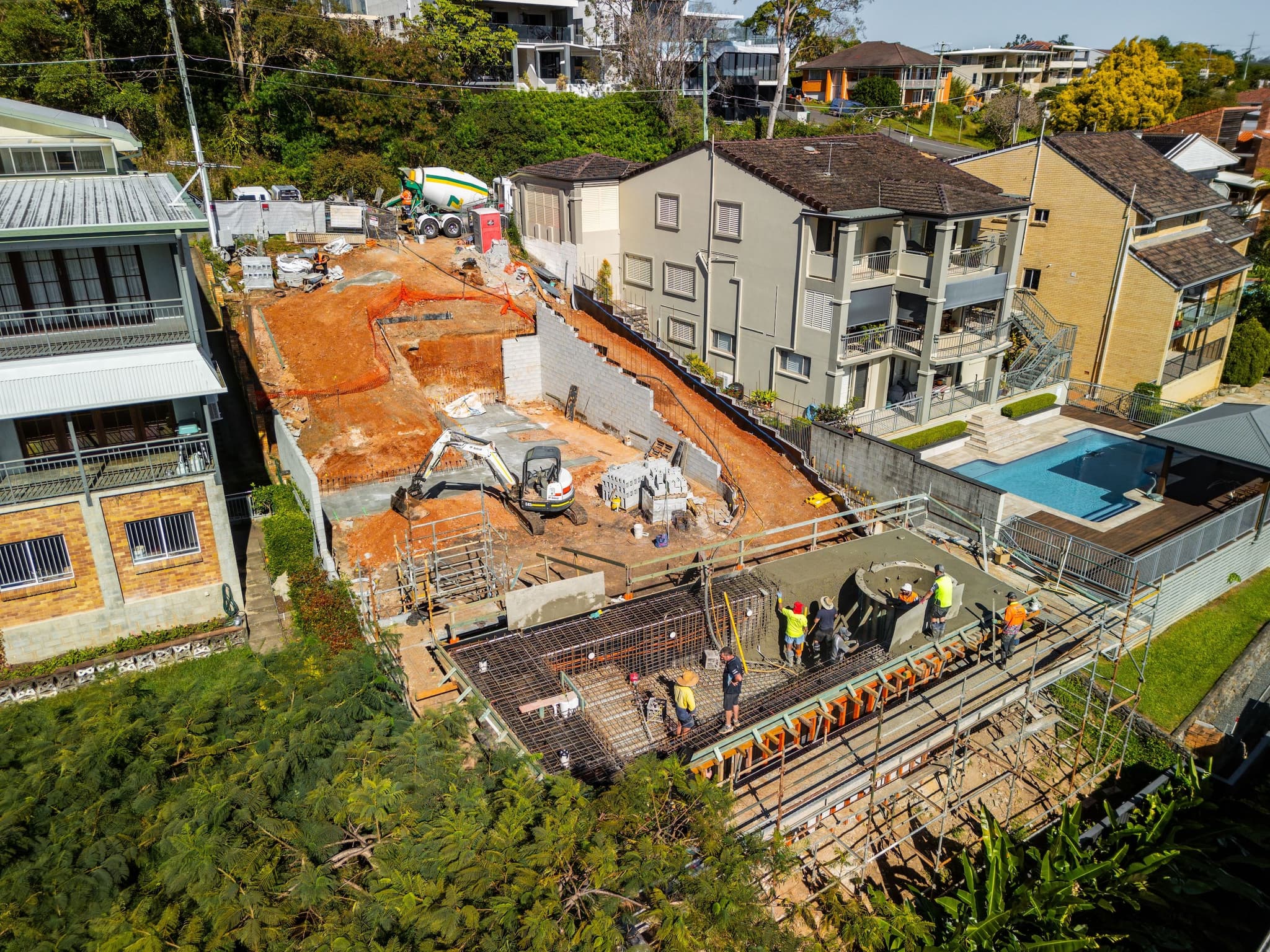 Aerial view of a sloping Brisbane block mid-construction, showing a concrete block retaining wall built into the slope, excavated site levels, and a reinforced concrete slab being poured by the site crew