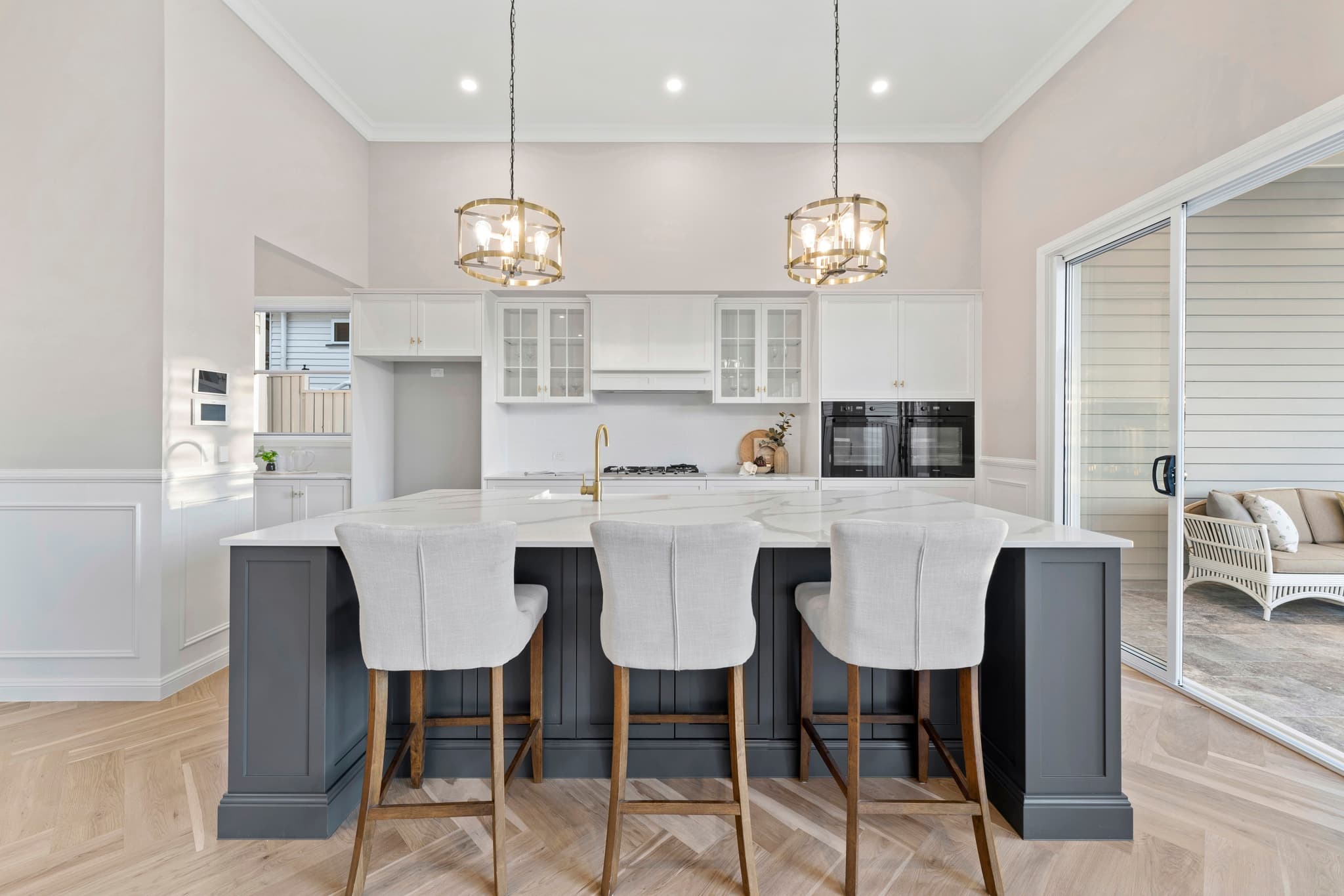 A custom luxury house design in Brisbane featuring herringbone floors, a shaker-style kitchen with a dark island bench, brass accents, and alfresco sliding doors.