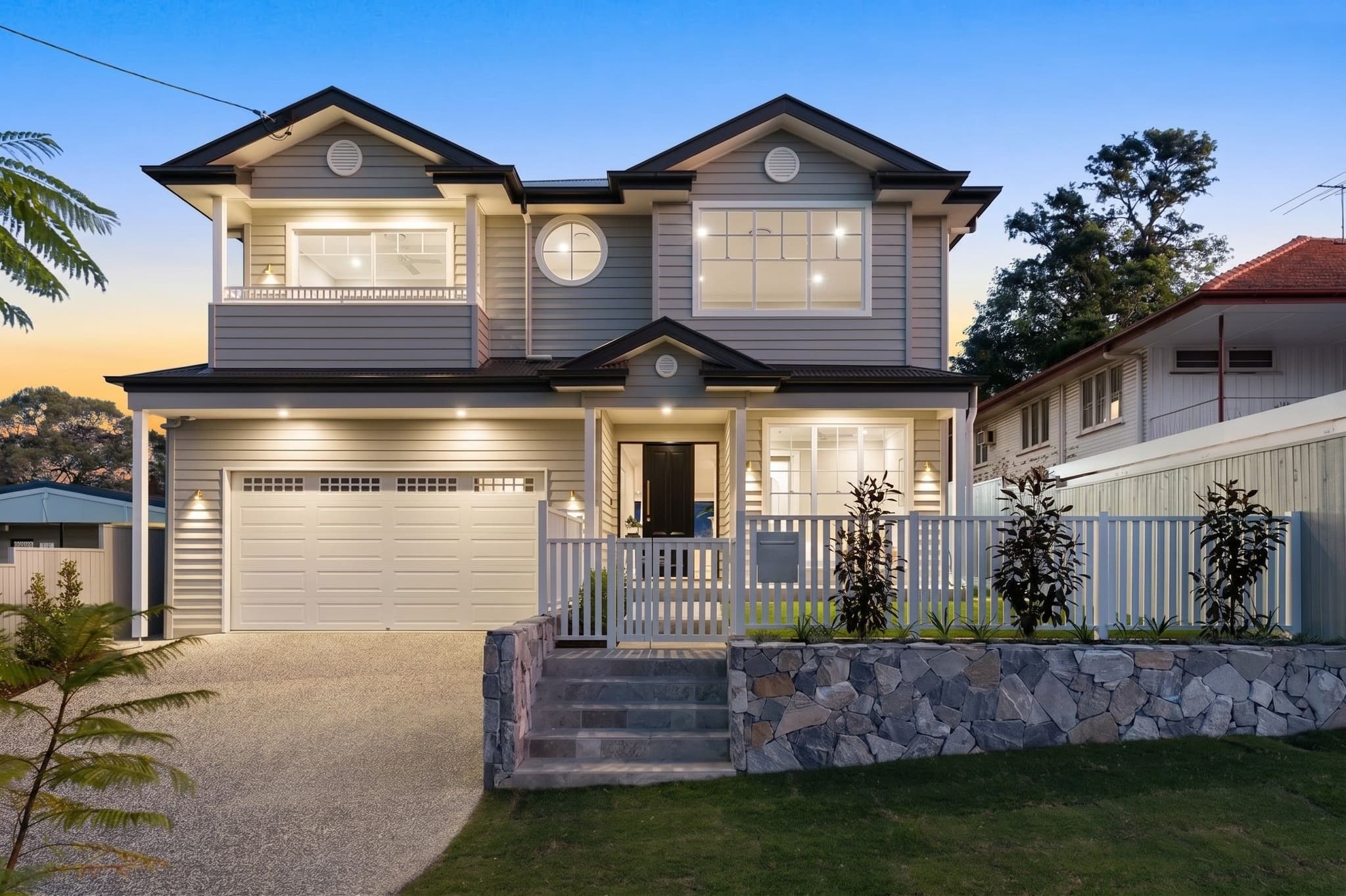 Hamptons style custom home facade at dusk in Balmoral, with weatherboard cladding, gable roofs and a stone retaining wall.